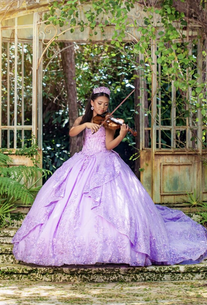 Quinceañera in a lilac ball gown playing violin in a rustic garden during a The Barn 305 photoshoot.